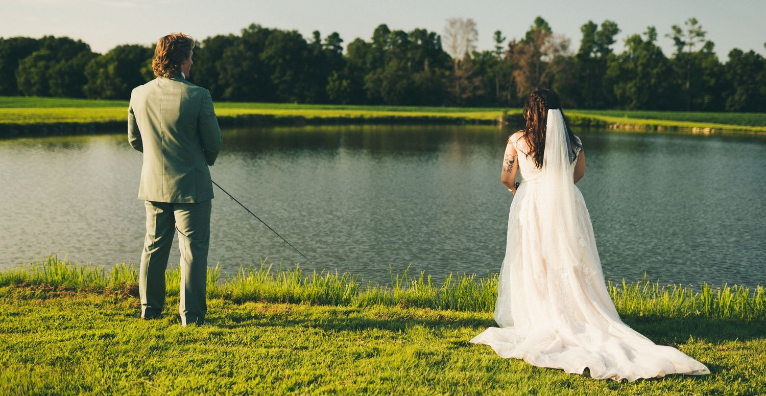 The bride and groom stand together at a lake at their venue, fishing, one of their favorite pastimes.