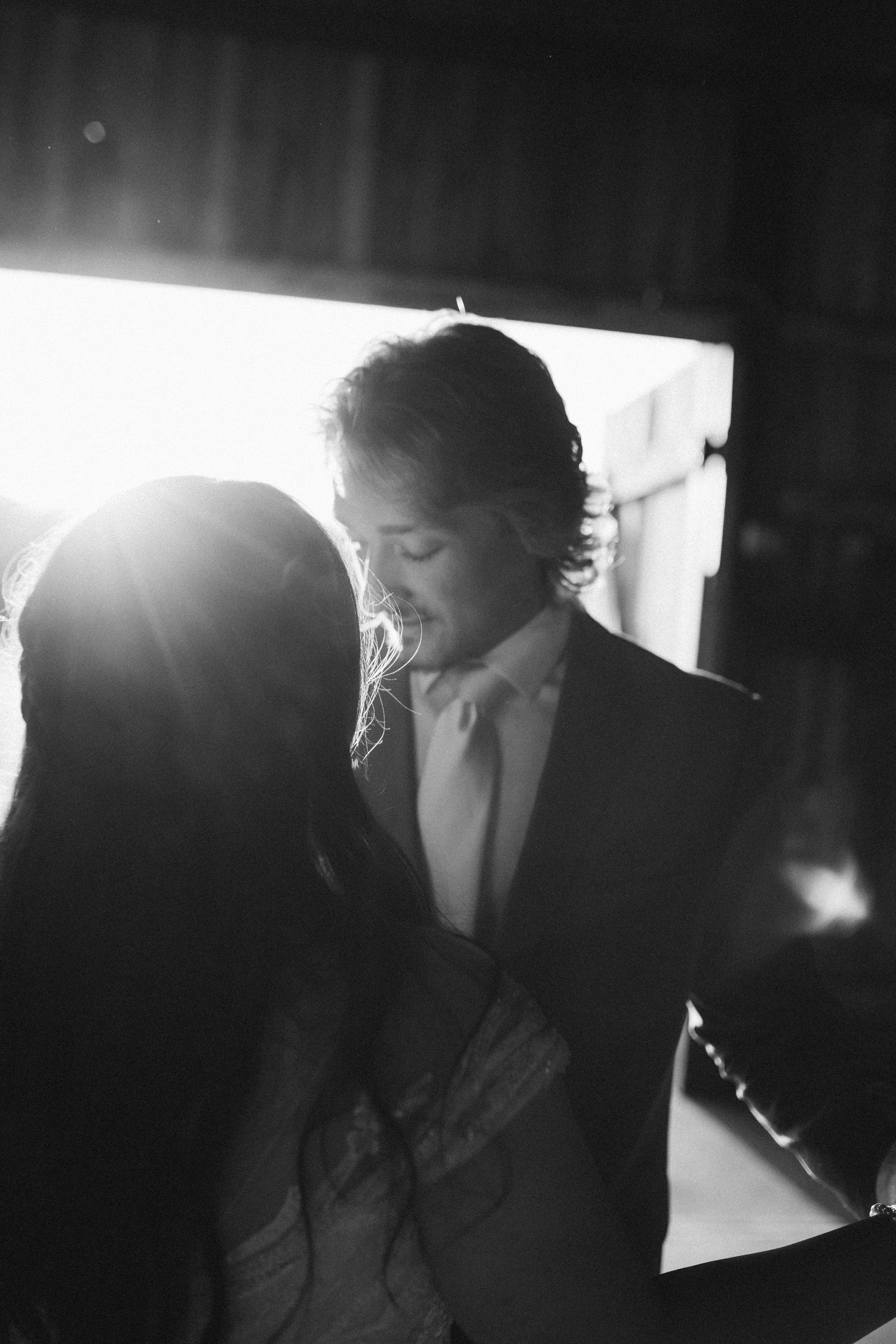 A closeup black and white photo of the bride and groom sharing their first dance. The golden hour sun makes an appearance above the bride.