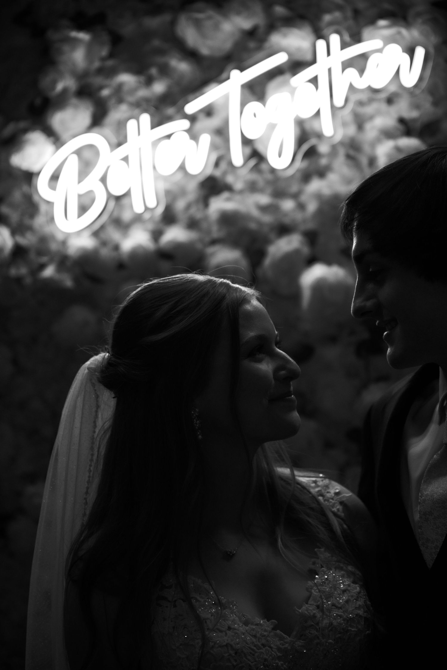 A slight silhouette of the bride and groom standing in front of an LED sign that says Better Together. The photos is in black and white.