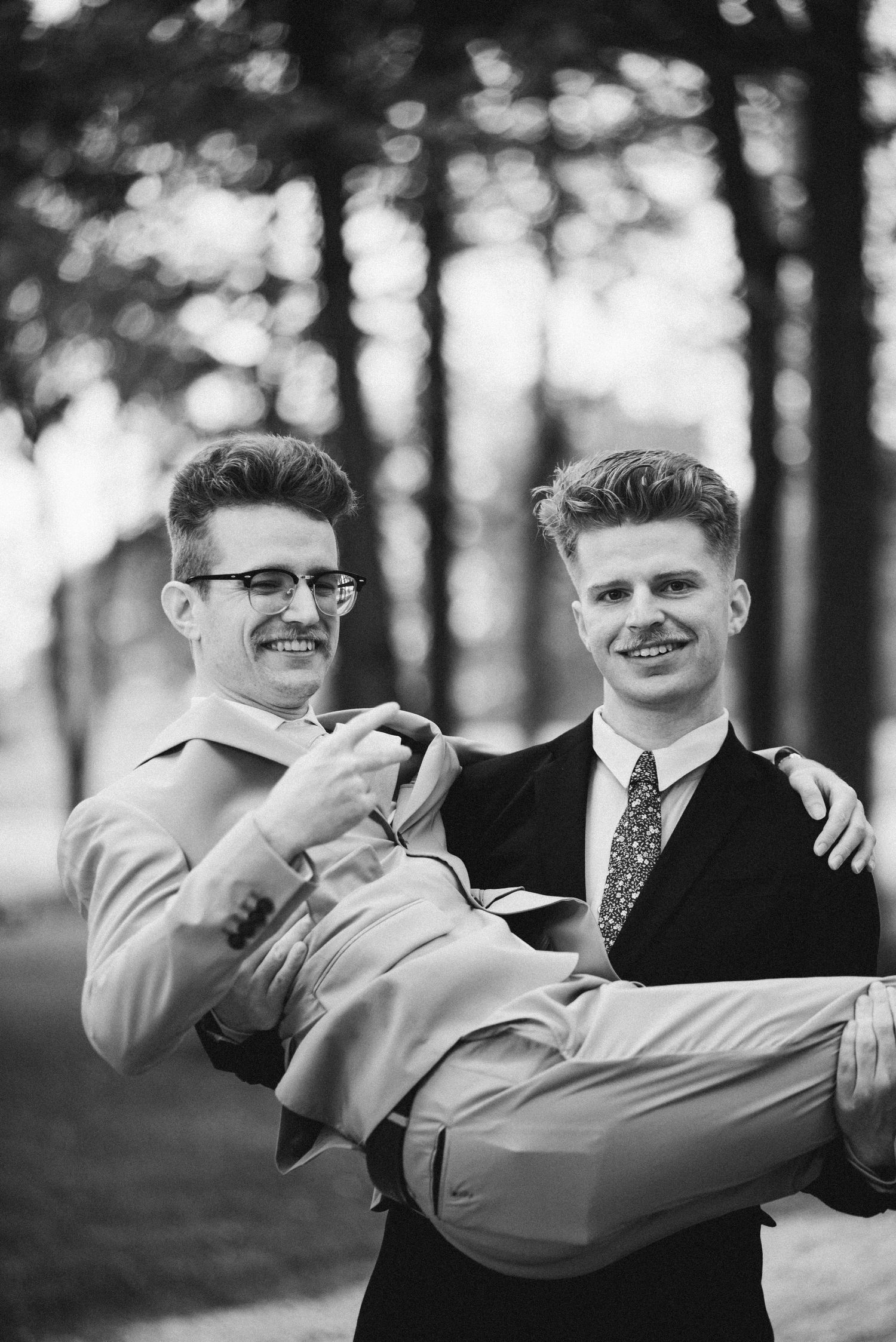 Myles laughs and holds up a peace sign as his best man picks him up and carries him bridal style. The photo is in black and white