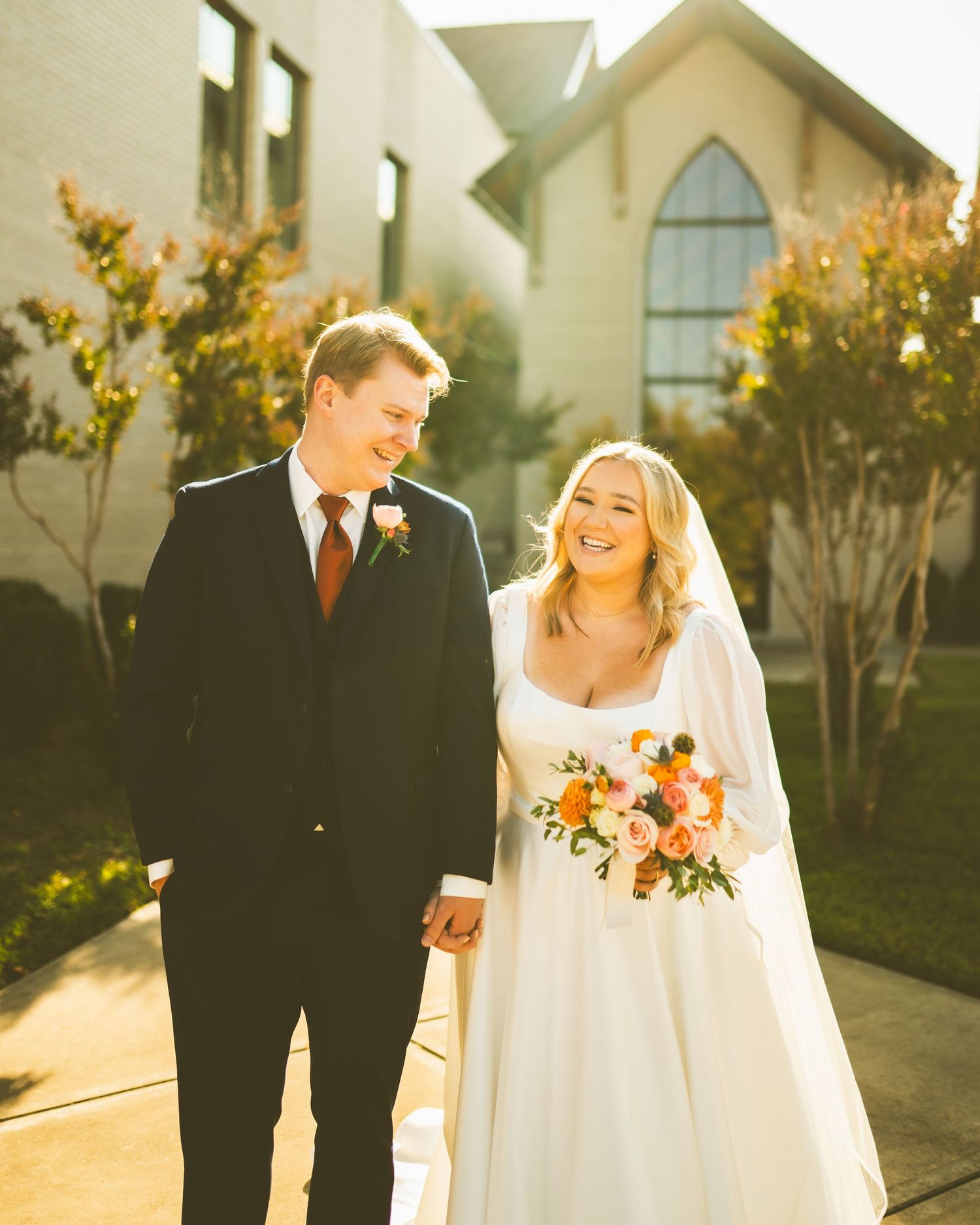 The bride, Sally, and her groom hold hands and share a sincere laugh as they walk in front of the church they married in