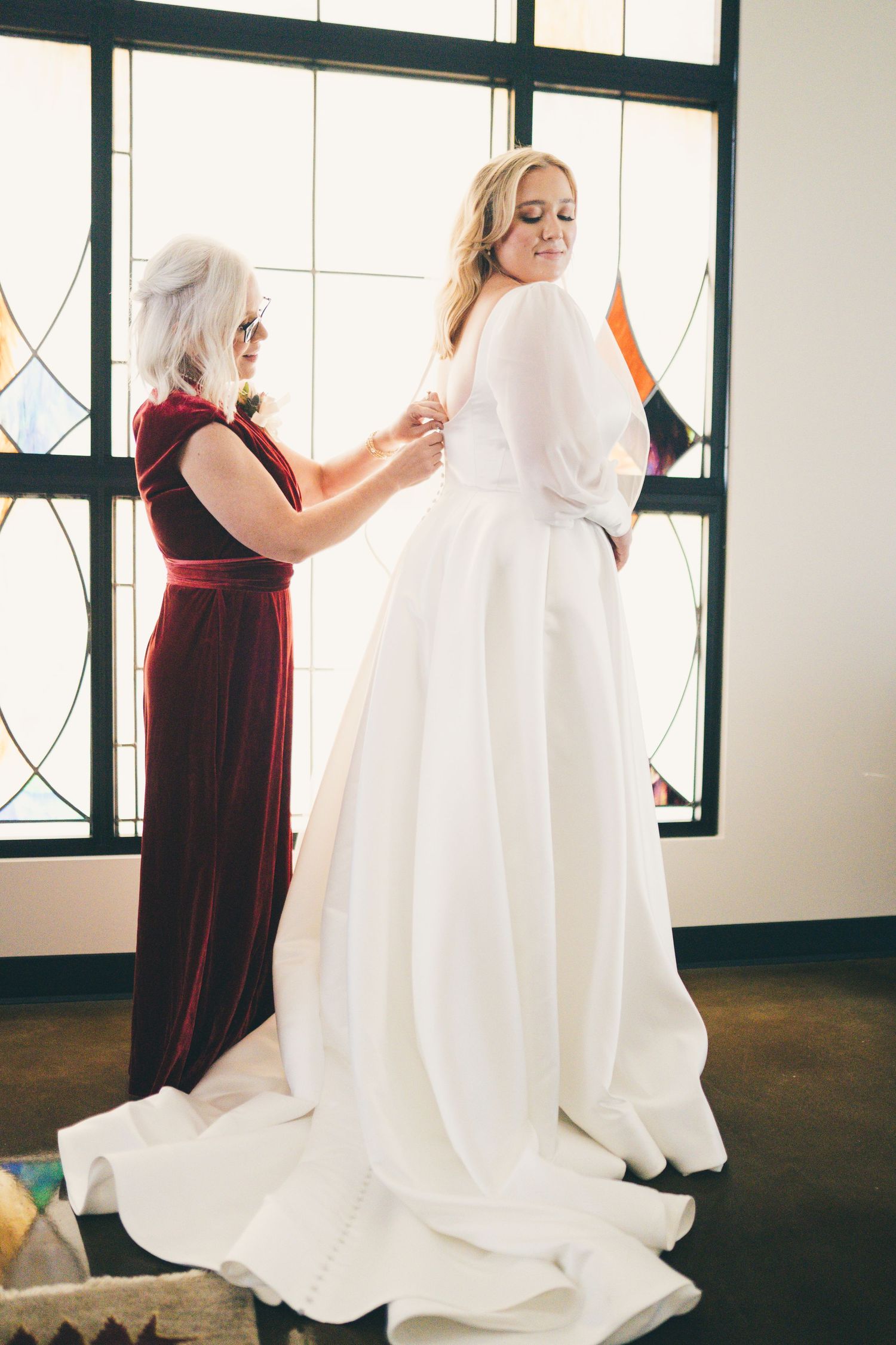 The bride & her mother share soft smiles, standing in front of the stained glass windows of the First Christian Church, buttoning her dress.