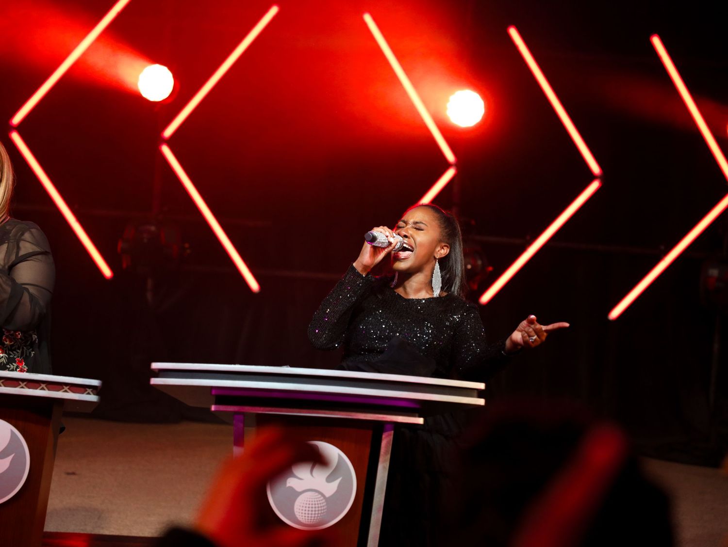 A performer in a sparkly black outfit sings on stage with dramatic red lighting and geometric shapes in the background.