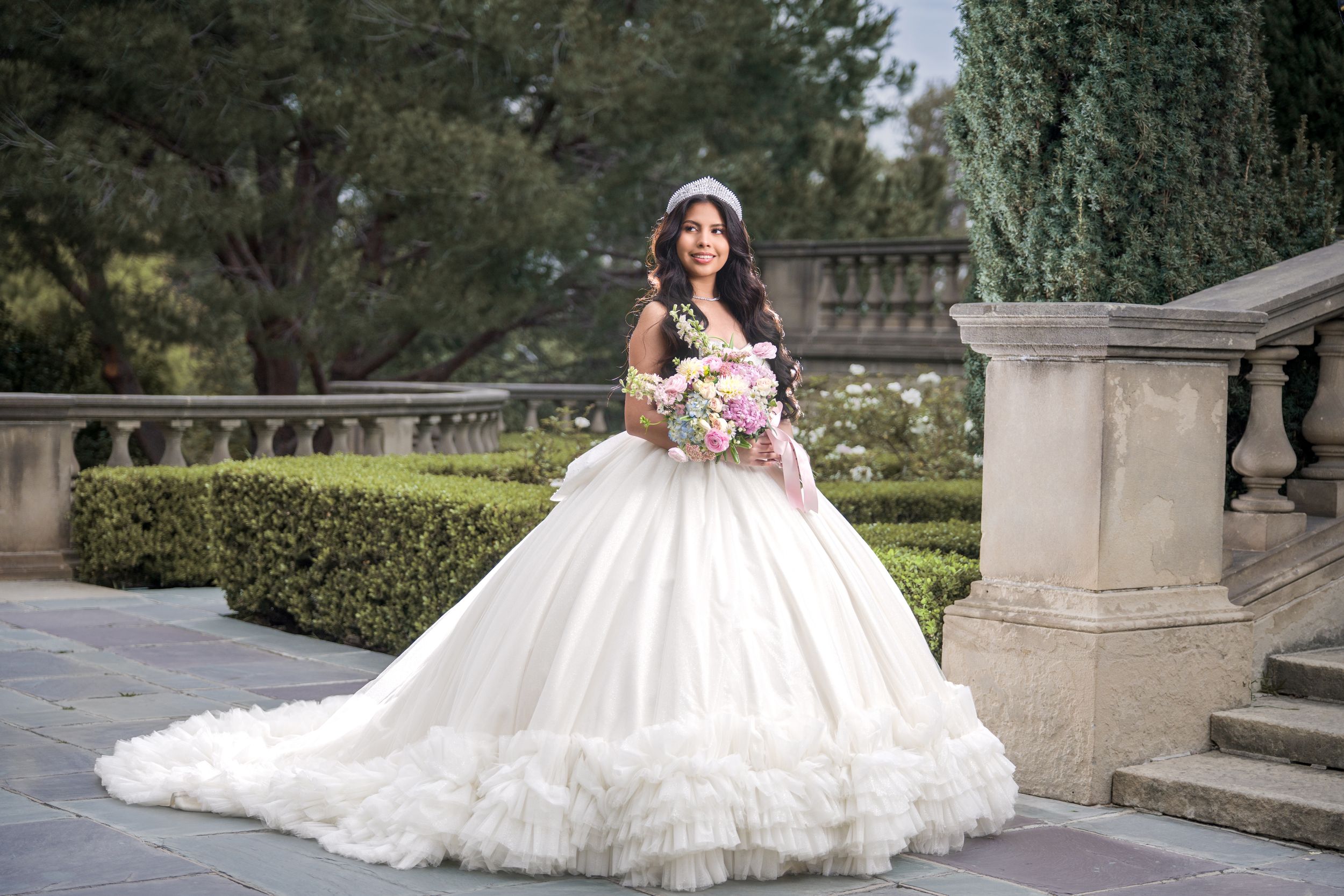 A stunning white ball gown wedding dress with a flowing train photographed on elegant stone steps surrounded by greenery.