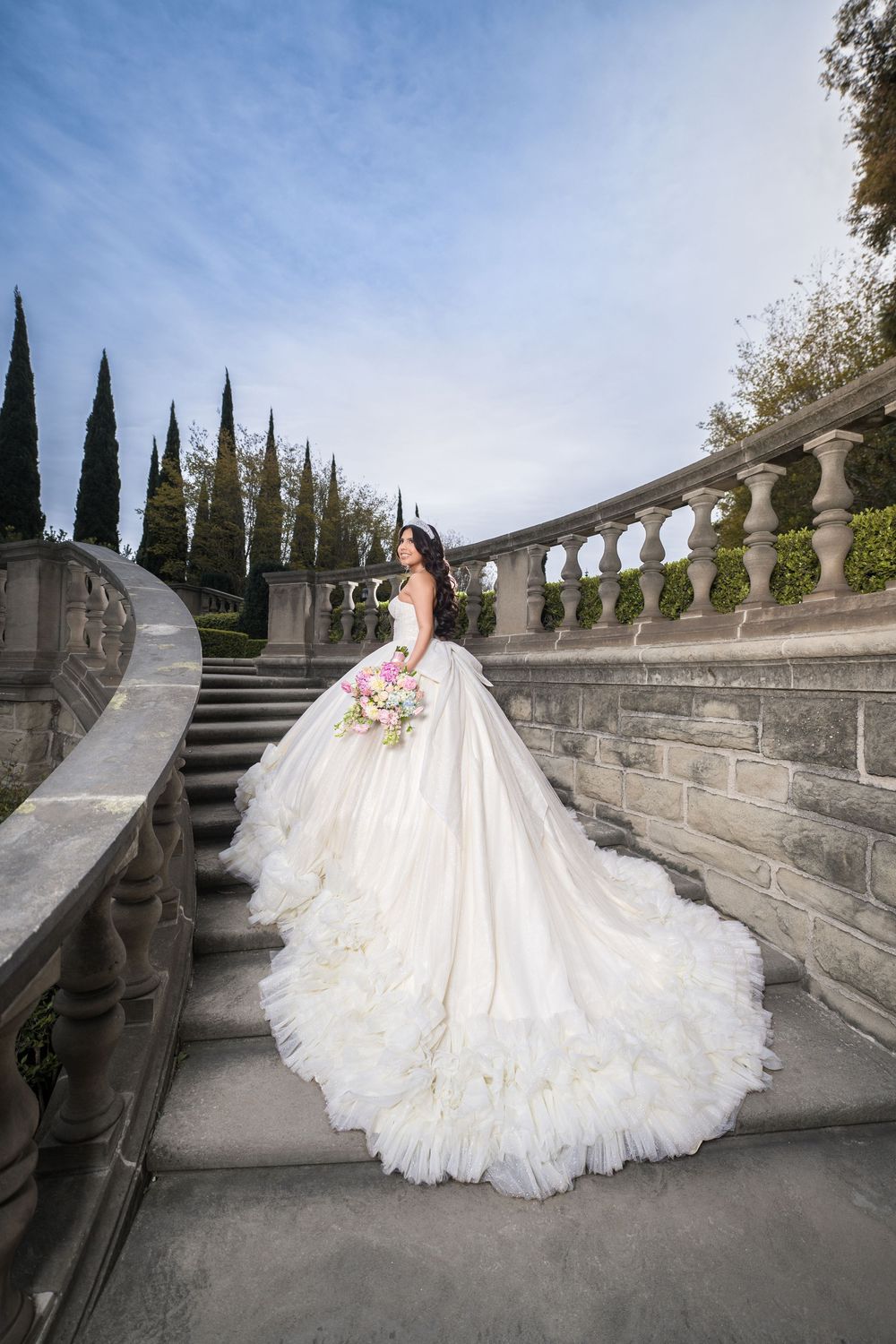 A flowing white wedding gown with a dramatic train cascades down elegant stone steps lined with cypress trees.