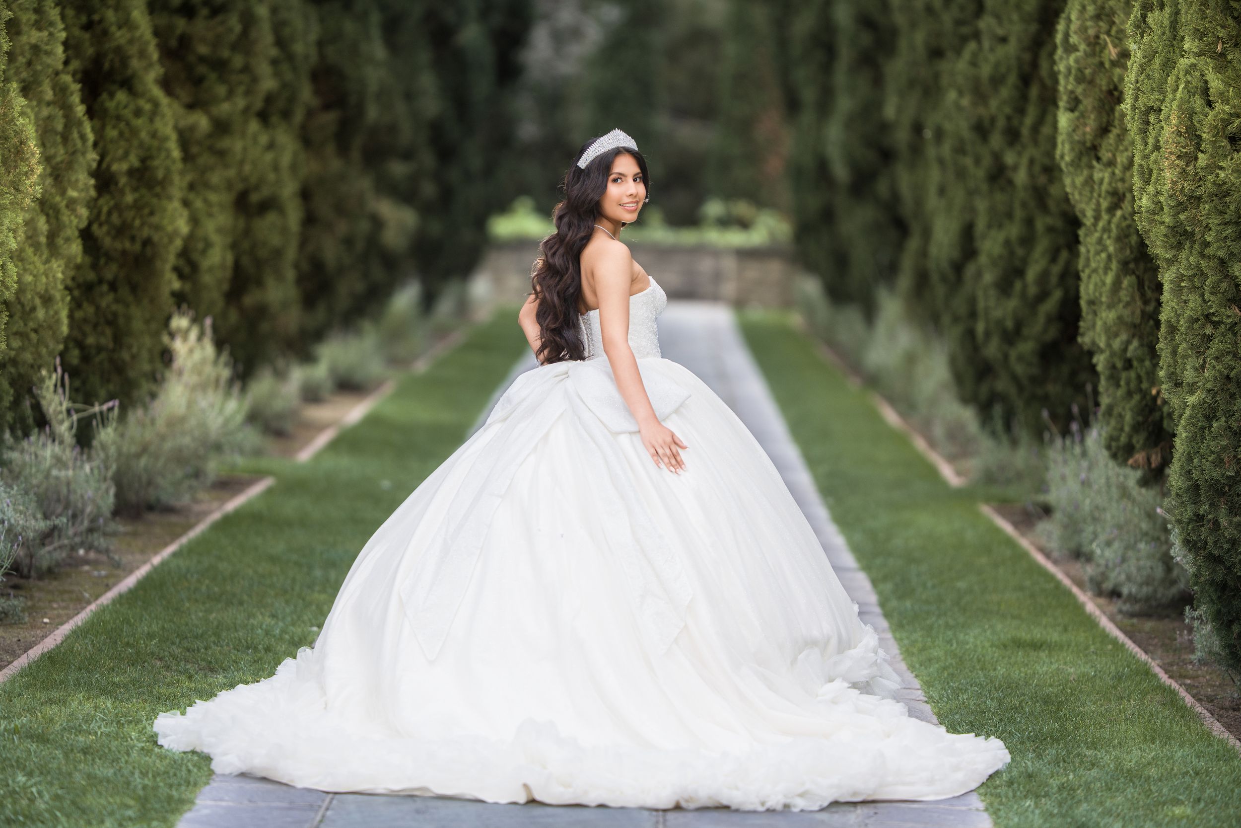 A bride in a flowing white wedding gown poses gracefully along a tree-lined garden path.