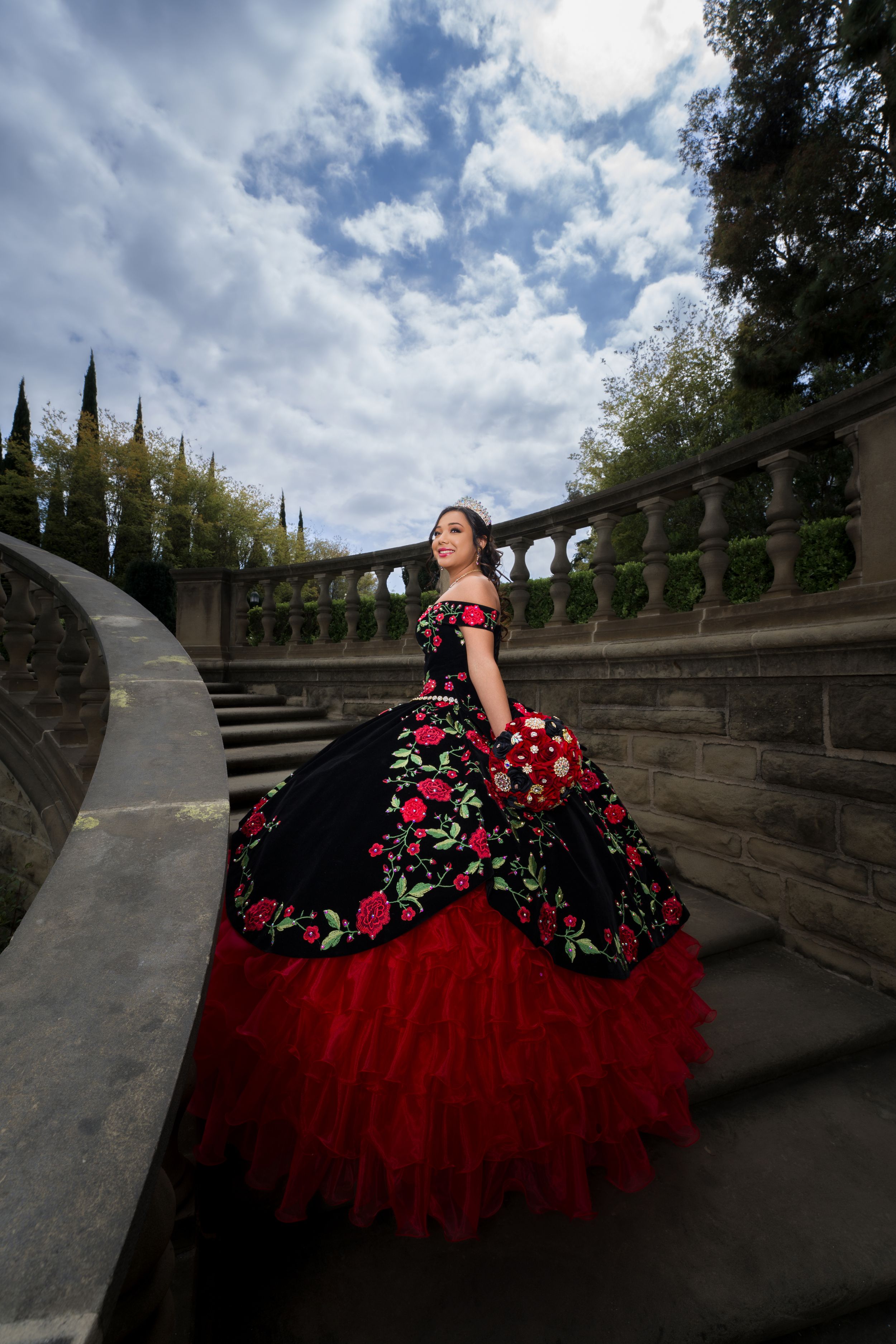 Brilliant red and black floral quinceañera dress photographed at dramatic angles on stone steps under cloudy skies.