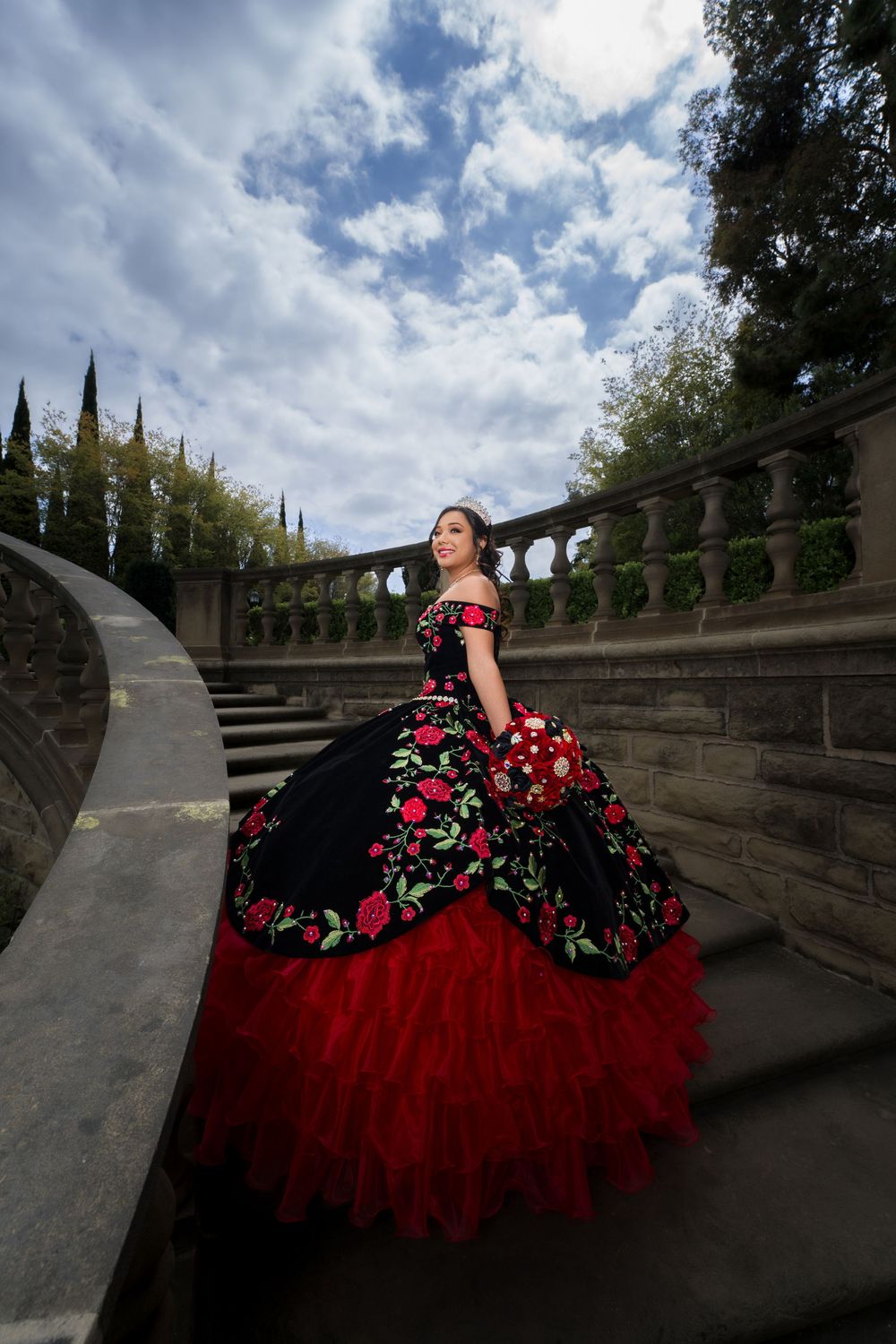 Brilliant red and black floral quinceañera dress photographed at dramatic angles on stone steps under cloudy skies.
