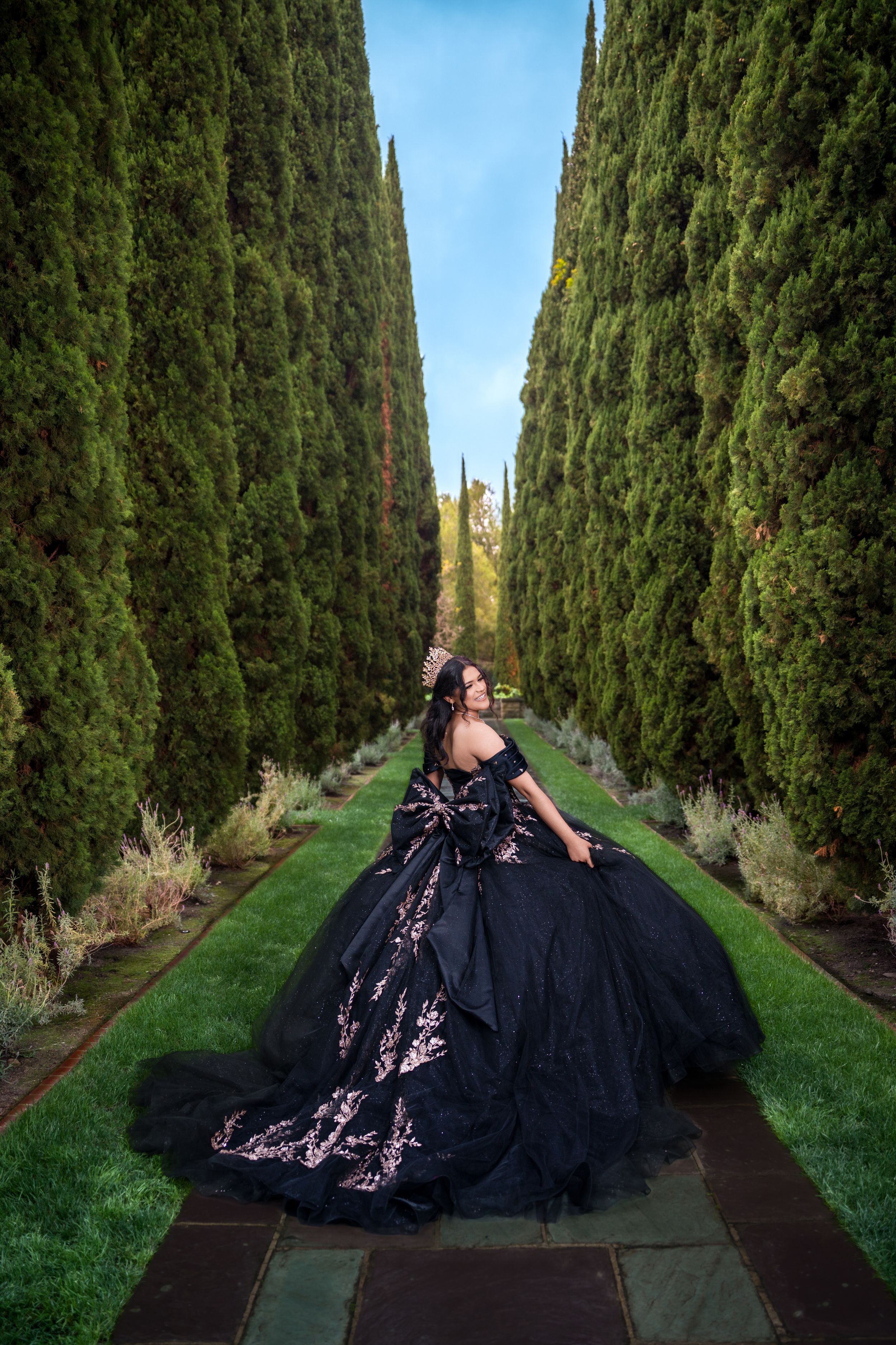 A figure in a dramatic black ballgown poses between tall cypress trees on a garden path under blue sky.