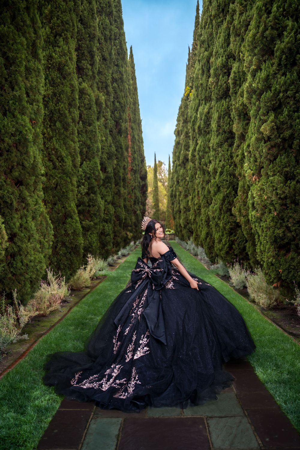 A figure in a dramatic black ballgown poses between tall cypress trees on a garden path under blue sky.