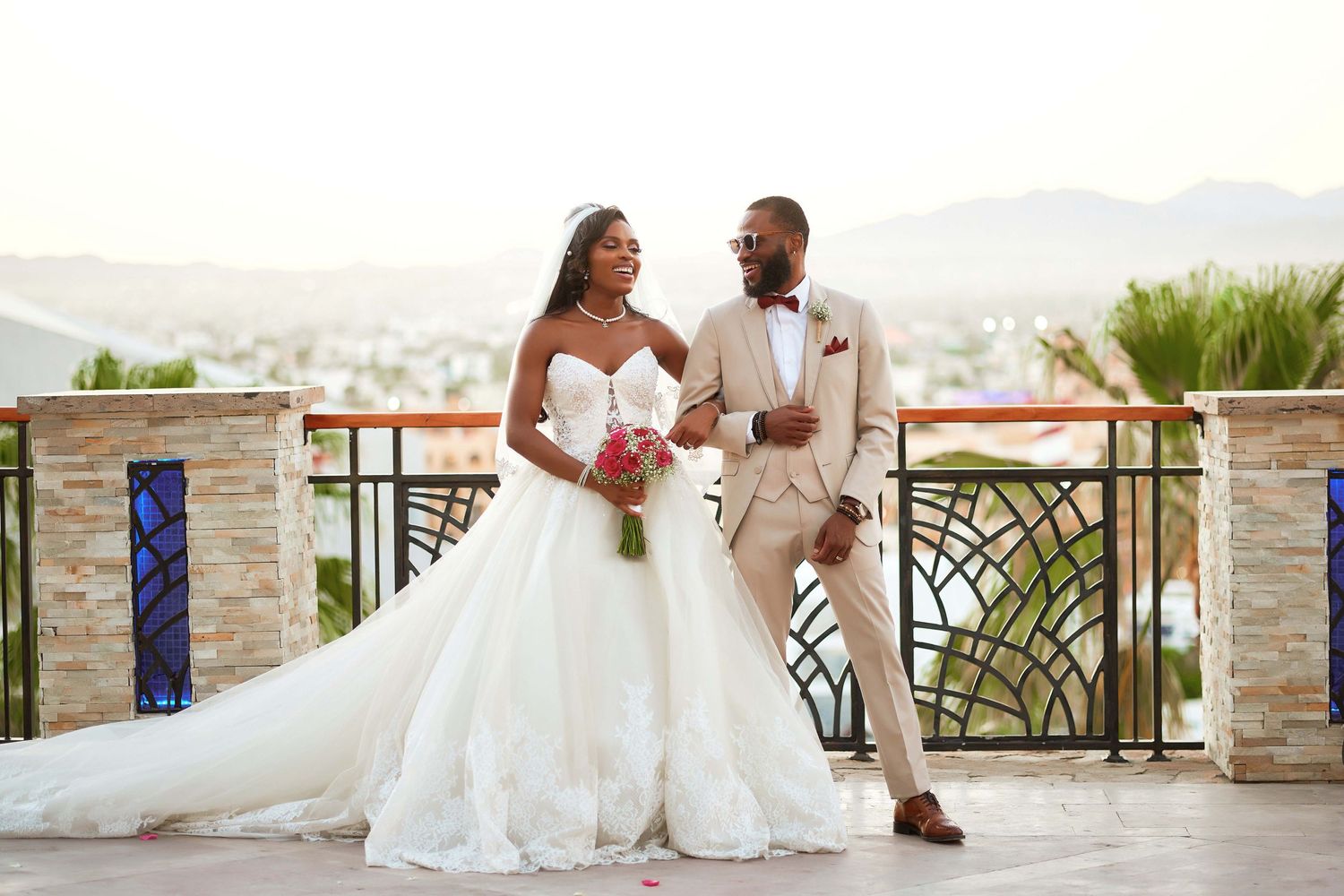 Newlyweds in formal wedding attire pose on balcony with mountain view backdrop.