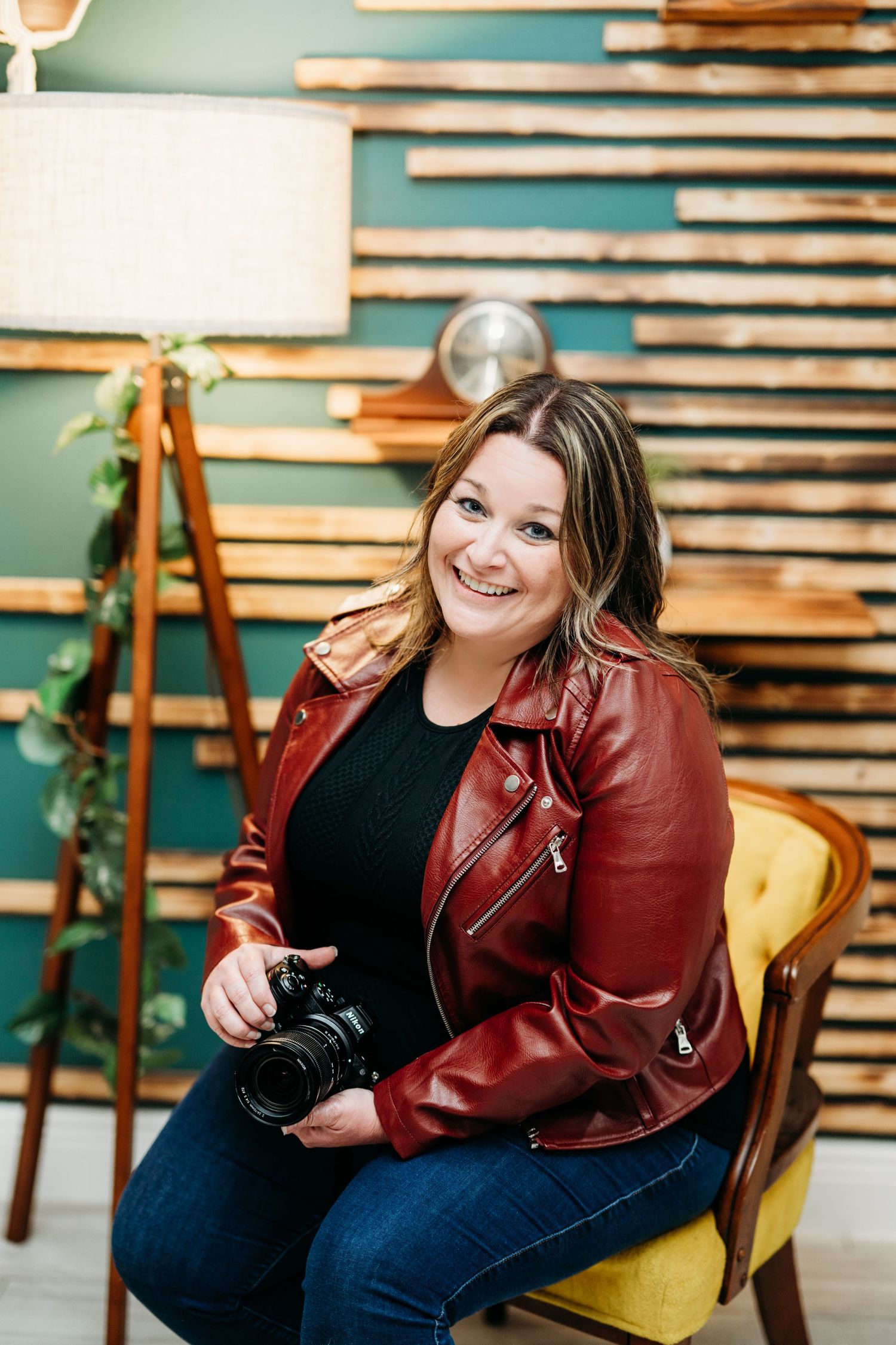 Photographer holding a camera while seated in yellow chair against wooden slat wall wearing red leather jacket.