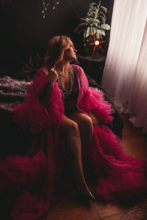 A person in a flowing hot pink tulle robe poses dramatically near a window in moody boudoir lighting.