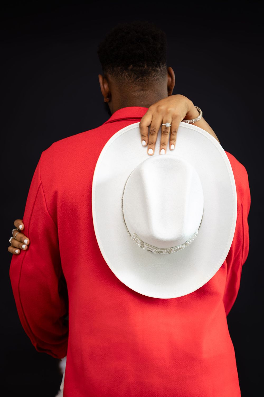 Back view of someone in a red jacket holding a white cowboy hat against a dark background.