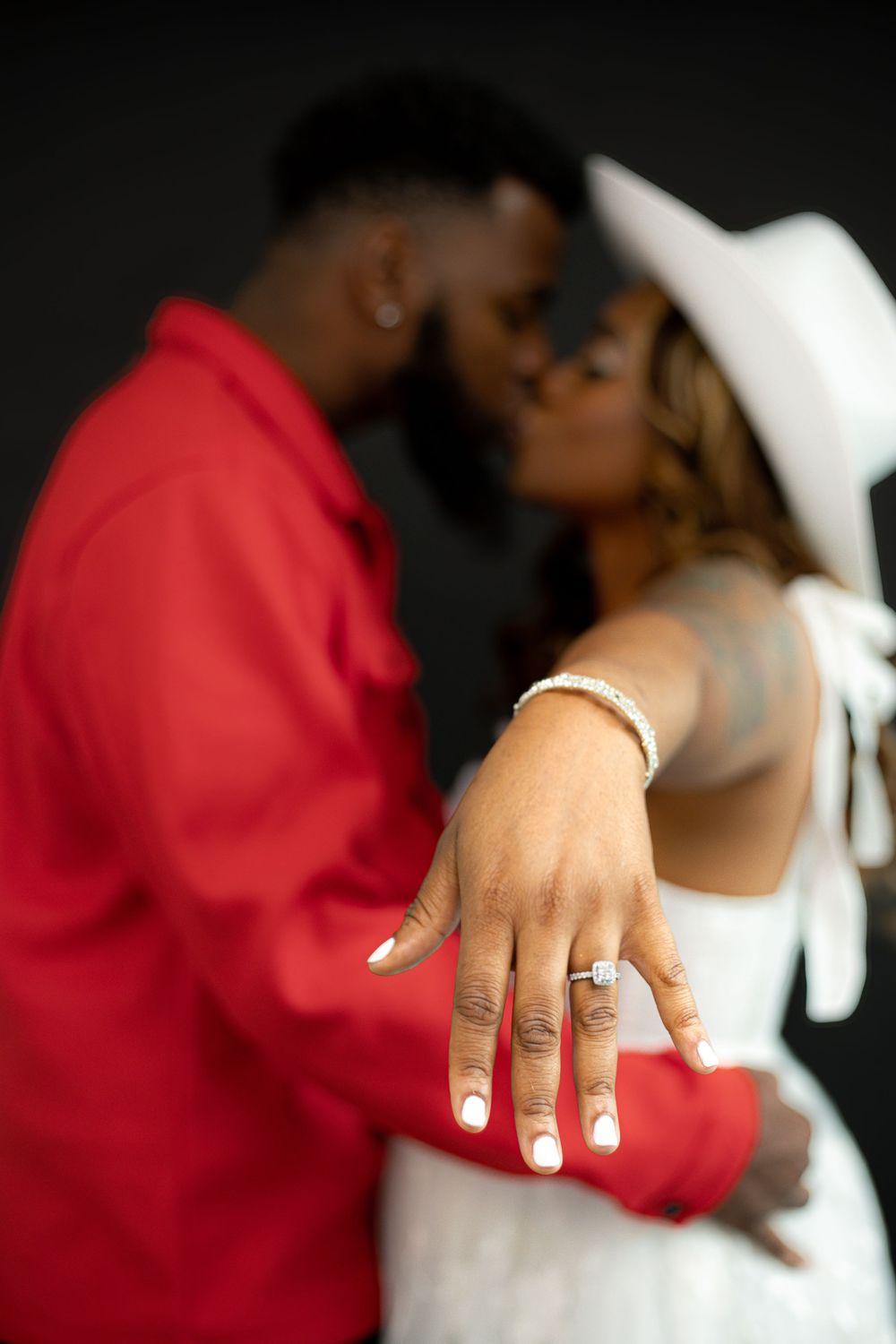 A close-up engagement photo showing a newly engaged hand with a diamond ring while a couple embraces in the background.