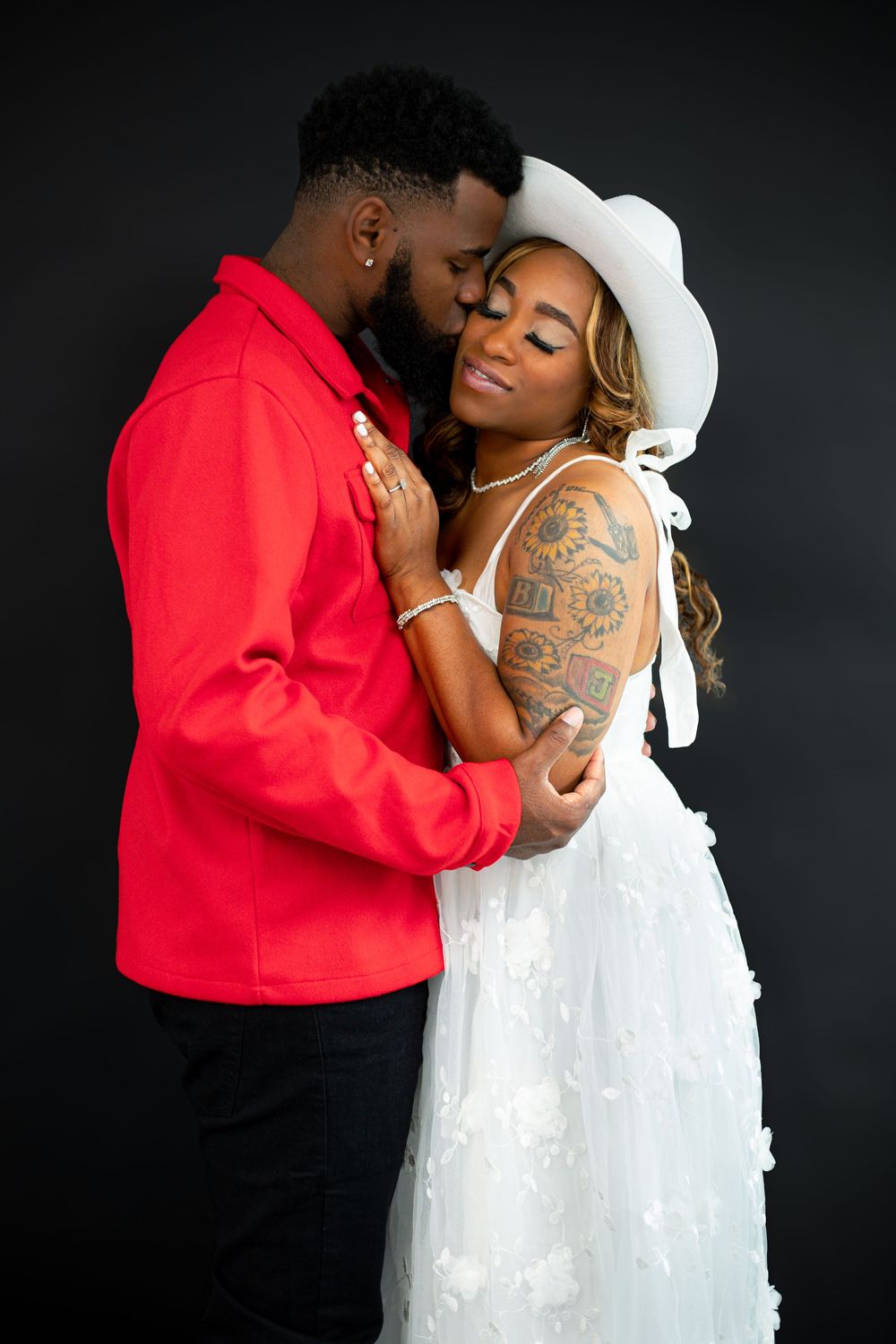 Romantic couple embraces against black background, with bride in white dress and groom in bright red jacket.