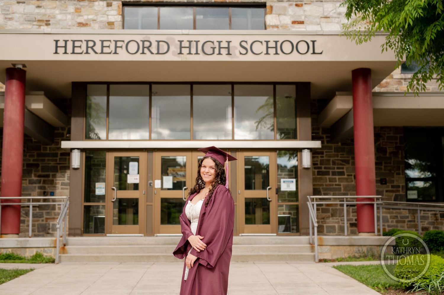 Graduate poses in burgundy graduation gown outside stone high school building entrance.