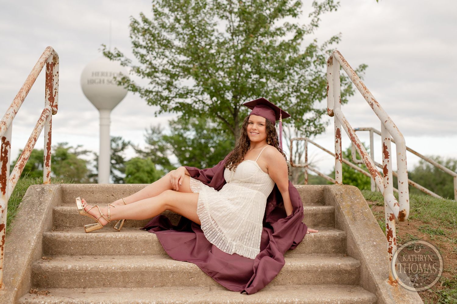 Graduate in white dress and maroon gown reclines on concrete steps with rustic railings.