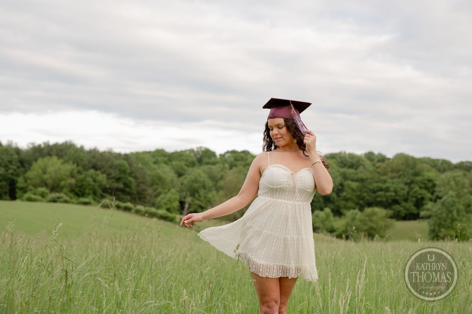 Graduate in white dress and maroon cap twirls joyfully in grass field.