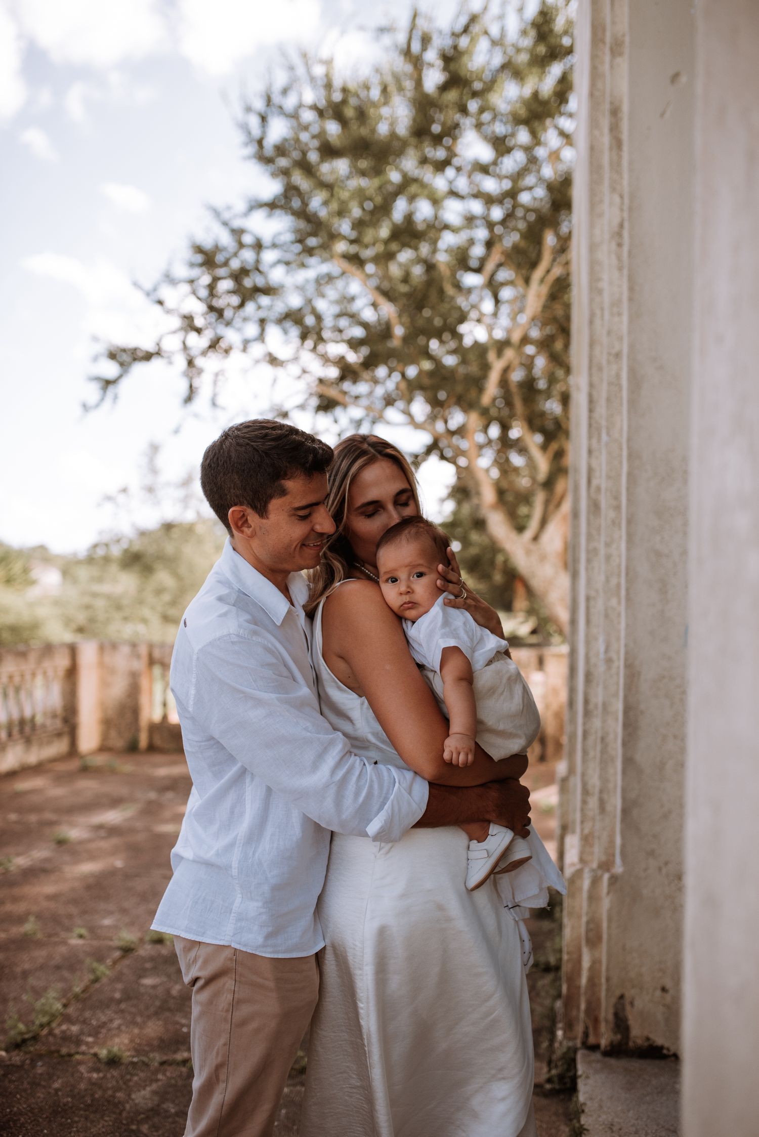A couple embraces while holding their baby outdoors near a white building.