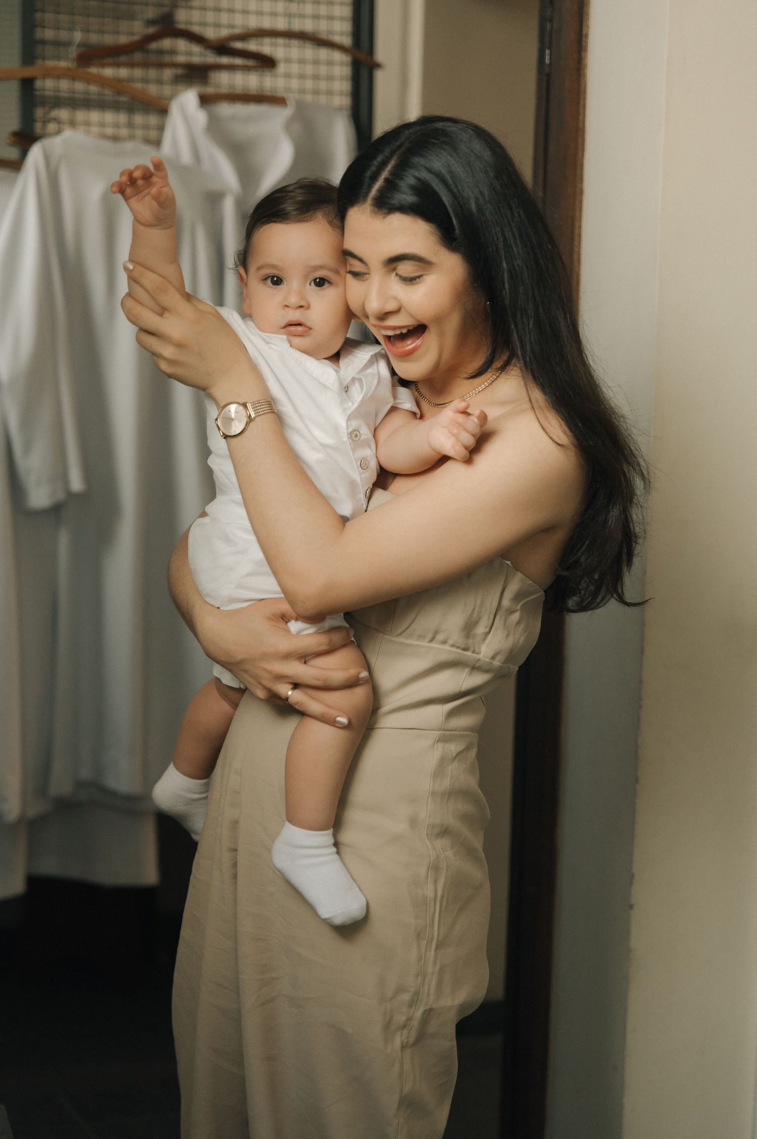 Mother in beige dress playfully holds laughing baby in white outfit.