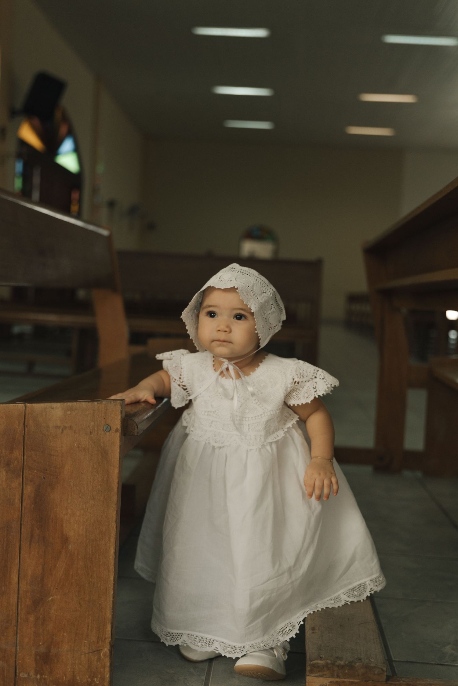 Baby in white baptismal dress and bonnet stands near wooden church pew.