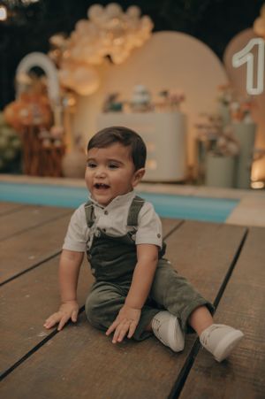 Happy toddler in overalls sitting on wooden deck at decorated party venue.