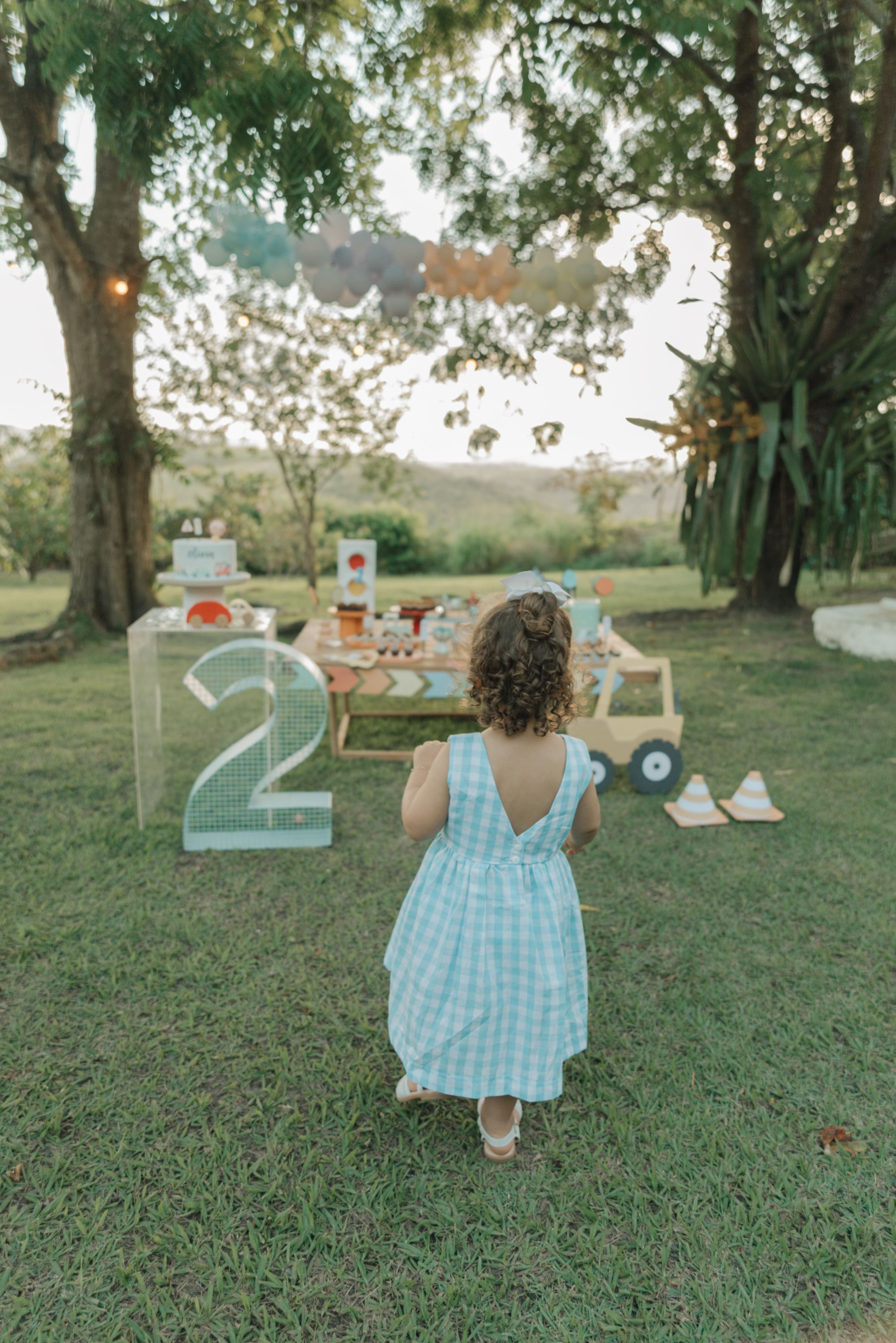 A child in a blue gingham dress plays at a whimsical garden party tea setup on a grassy lawn at sunset.