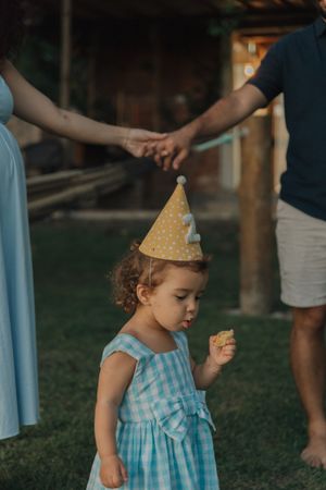 A small child in a blue gingham dress and party hat holds a cookie while walking on grass at sunset.