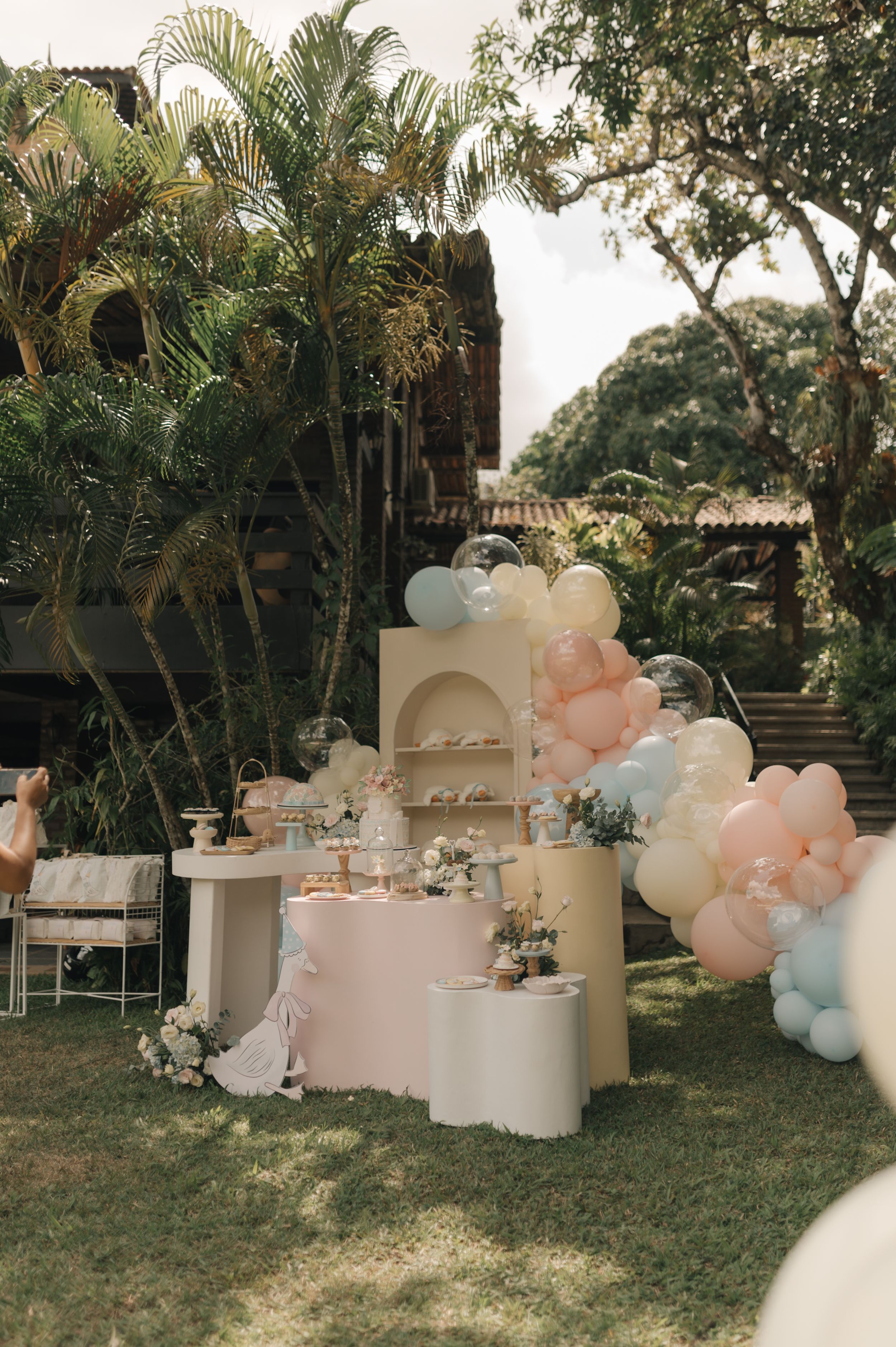 Elegant outdoor party setup with pink and white balloon decorations, tables, and palm trees creating a romantic garden atmosphere.