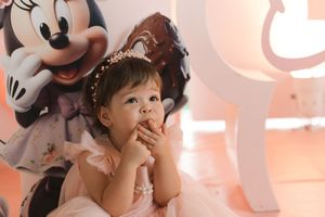 A child in a tulle dress poses thoughtfully against a pink backdrop with a Mickey Mouse balloon decoration.