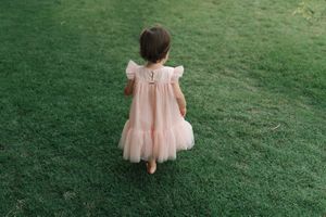 A small child wearing a pink ruffled dress stands alone on a grassy lawn viewed from behind.