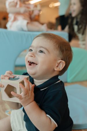 A happy baby in a dark sweater smiles and laughs while enjoying a drink from a disposable cup.