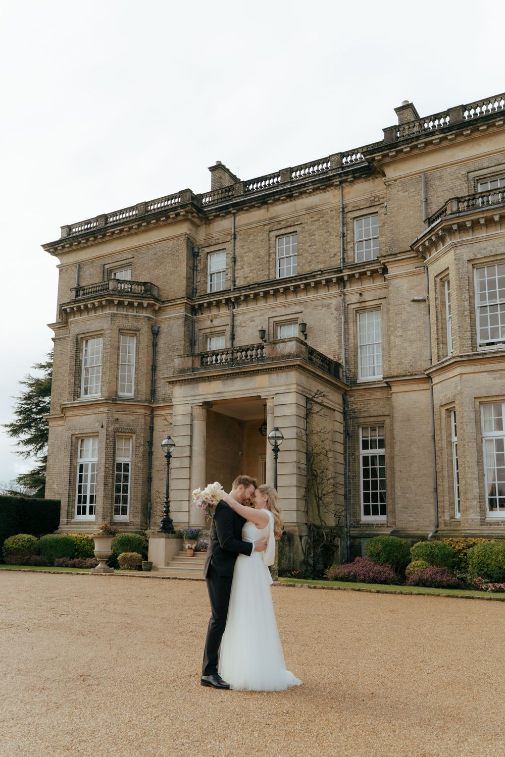 A couple shares a romantic moment outside a grand historic English manor house with ornate stonework and classical architecture.