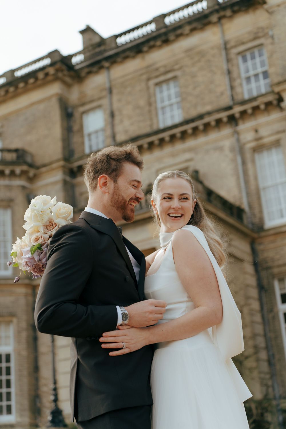 A joyful couple in wedding attire embrace and share a laugh outside a grand historic stone mansion.