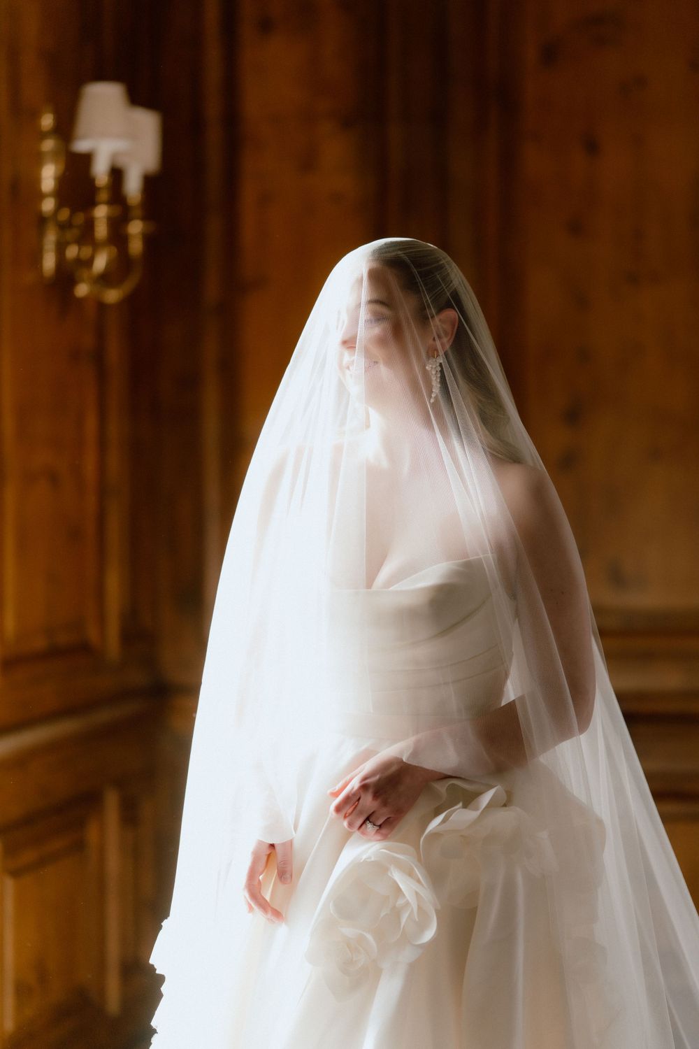Elegant bride in flowing white dress and delicate veil stands in warm wooden room with wall sconce lighting.