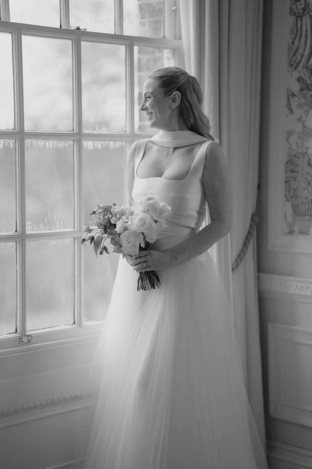 A black and white portrait of someone in a flowing white wedding gown holding a bouquet while standing by a window.