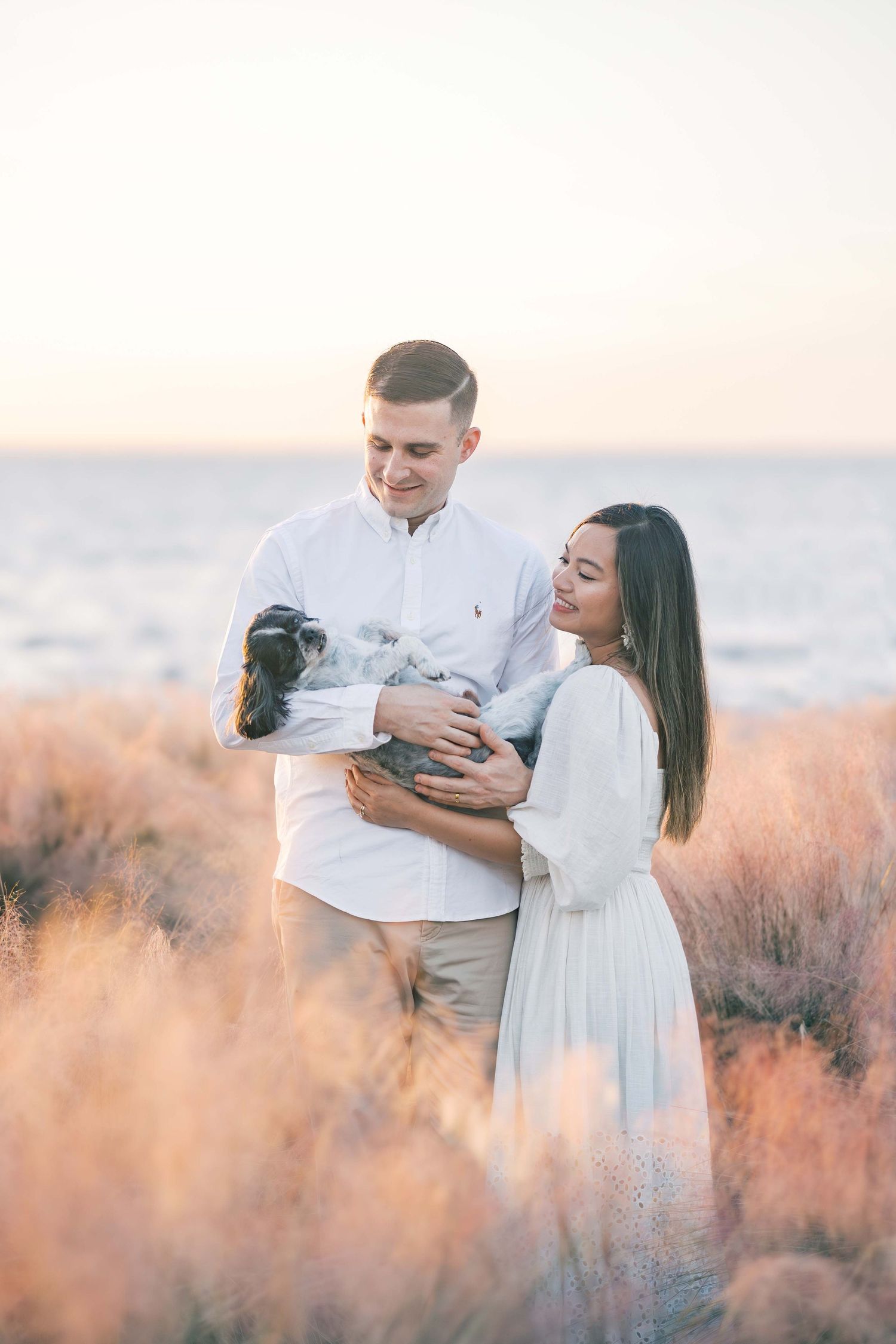 Couple embraces while holding their pet in field of tall grass at sunset.
