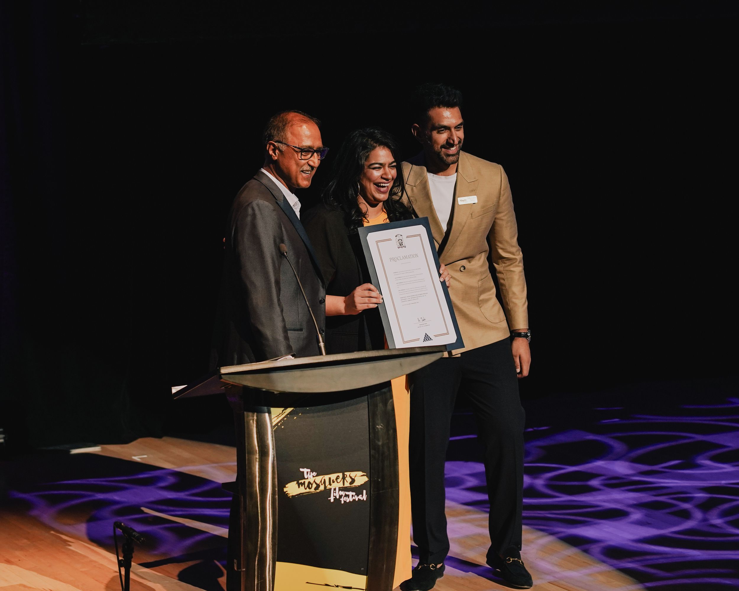 Three people stand at a podium on stage, holding a framed certificate during an award ceremony.