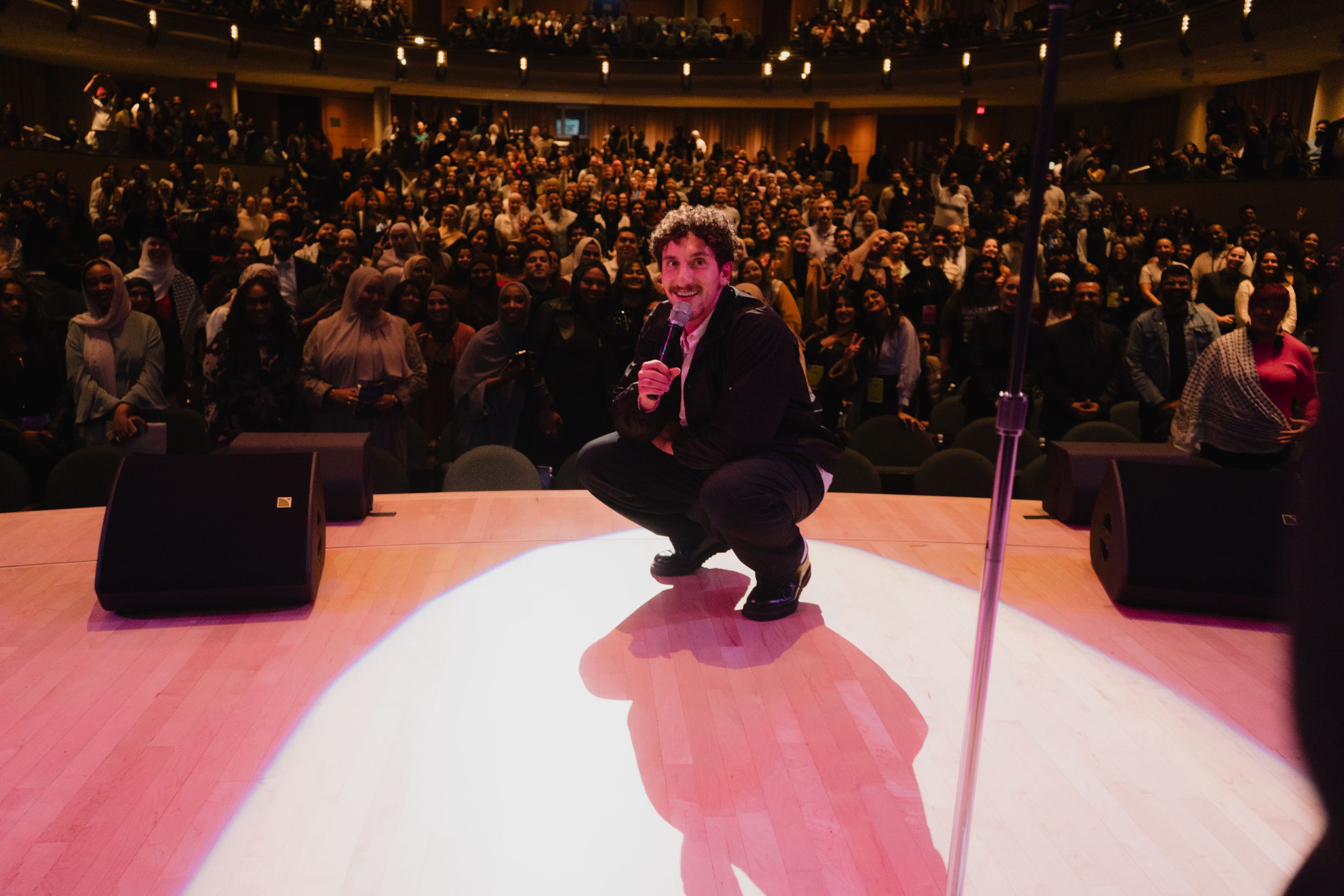 A performer crouches on a spotlight-lit stage in front of a large audience in a packed theater.