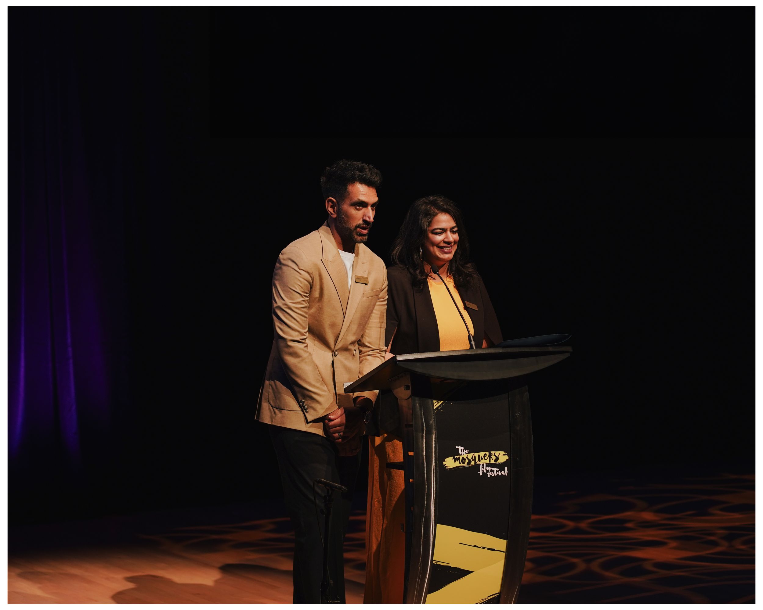 Two presenters, a man in a tan blazer and a woman in yellow, stand at a podium on a dimly lit stage.