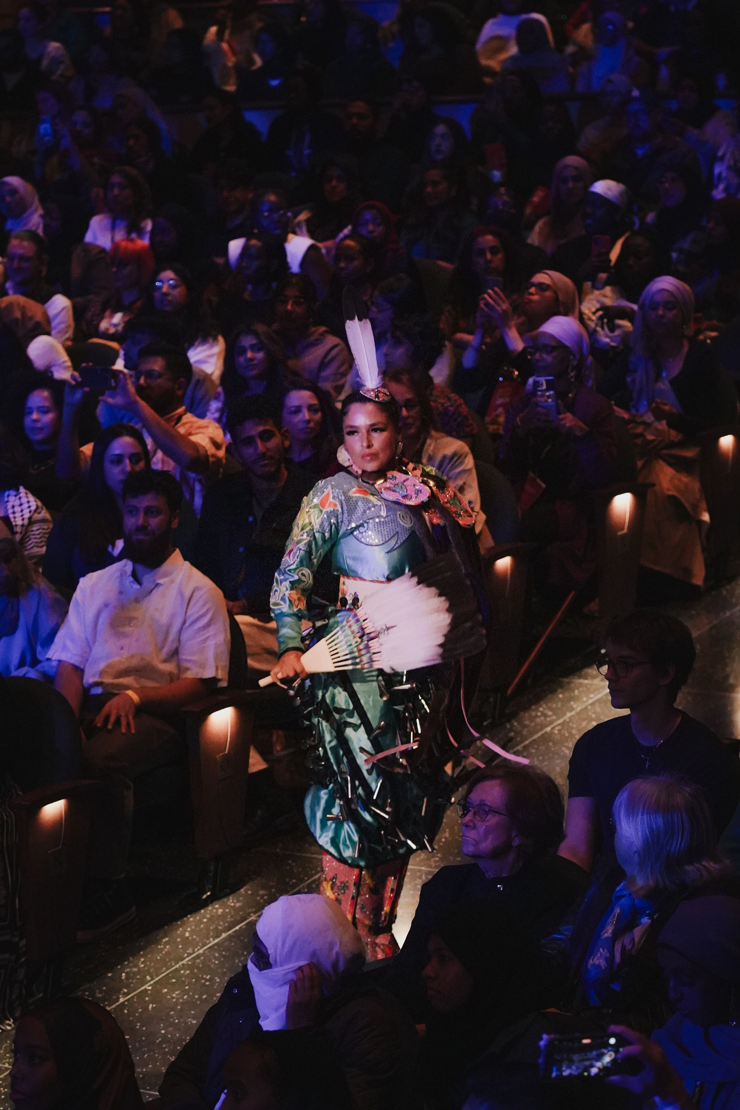 Performer in traditional costume walking through a dimly lit theater audience.