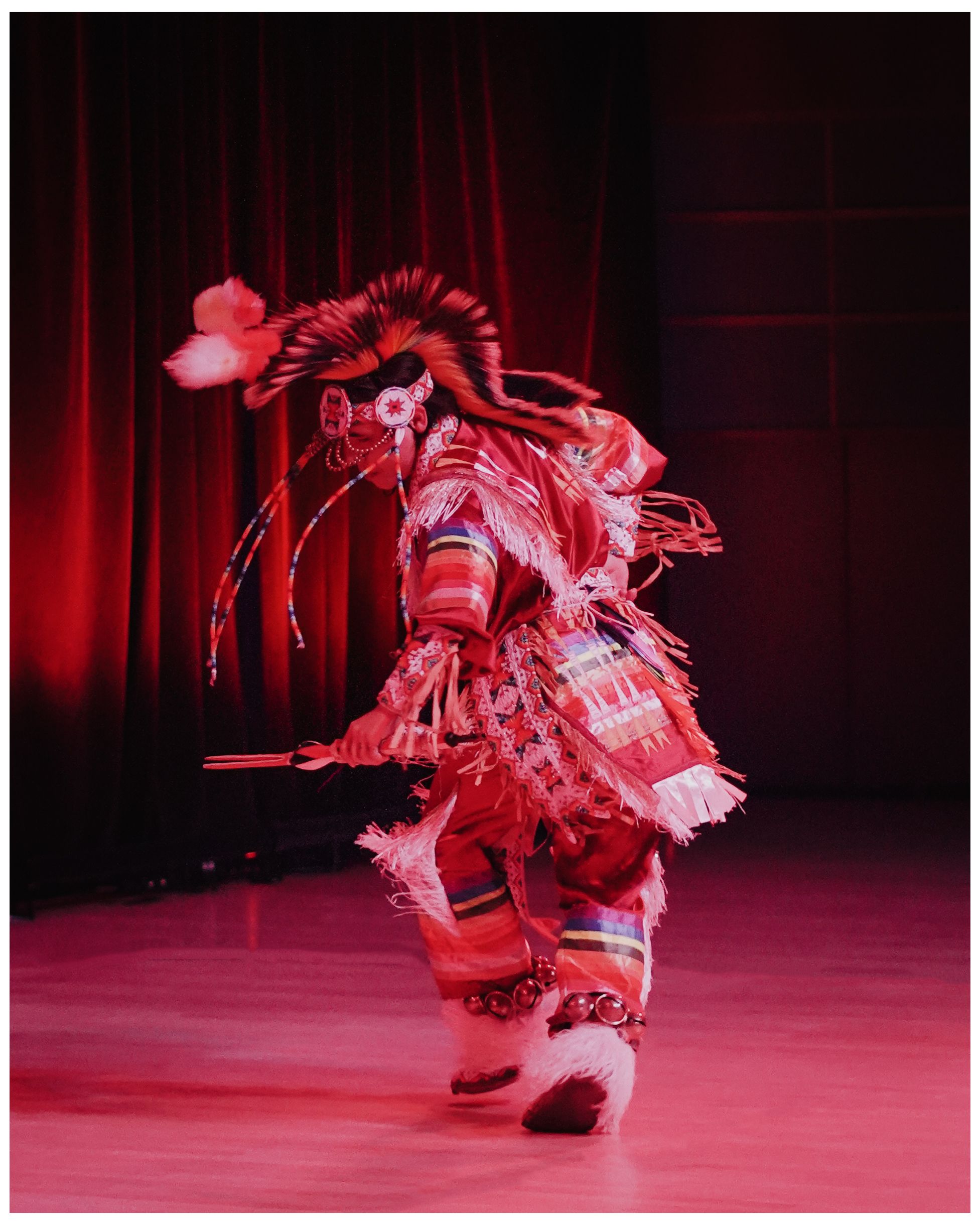 Traditional Native American hoop dancer performing on stage in red lighting.