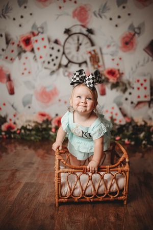 Baby in mint outfit sitting in vintage rattan furniture against floral backdrop.