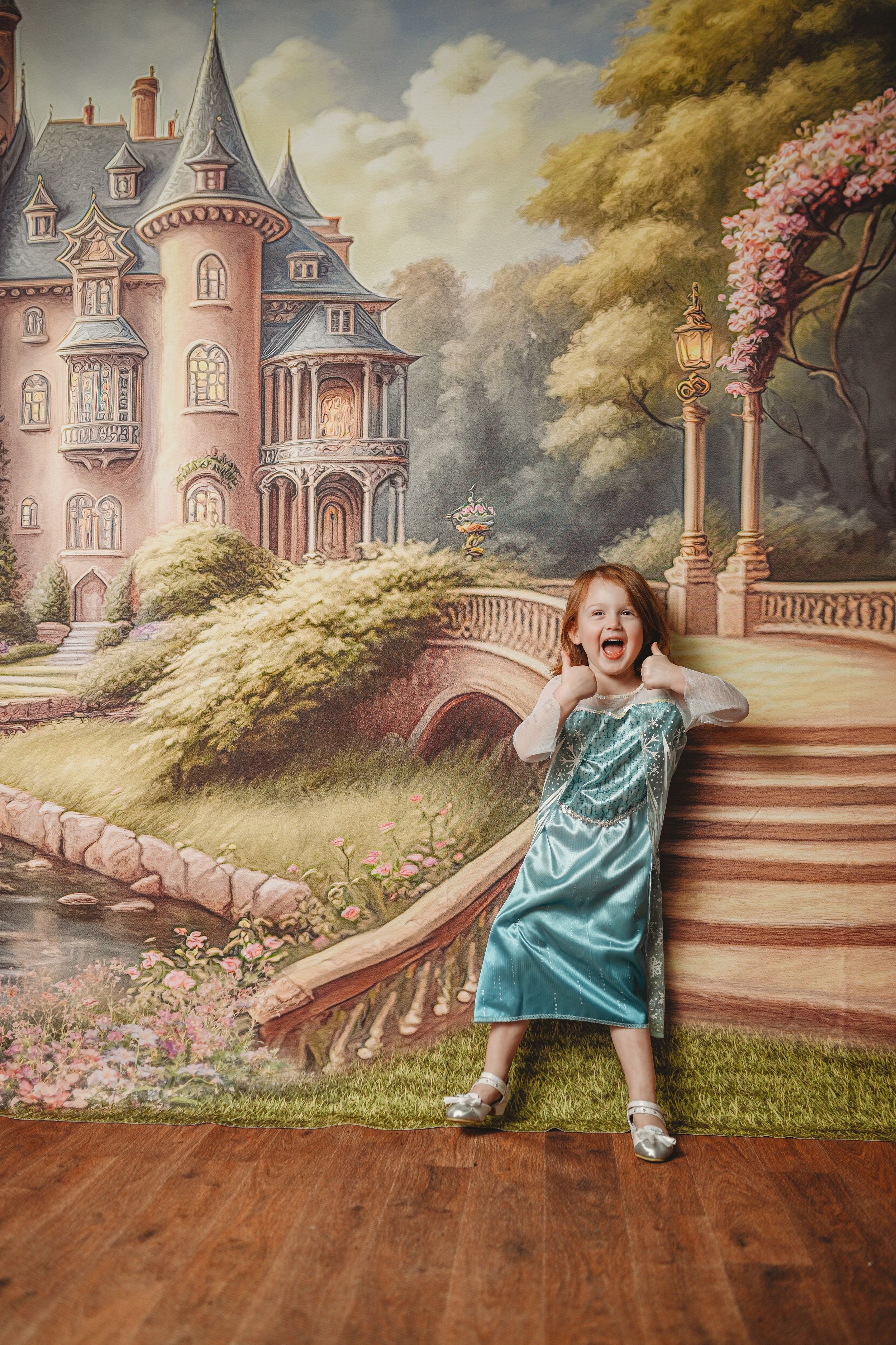 A joyful red-haired girl in a teal dress poses in front of a fairy tale castle painted backdrop.