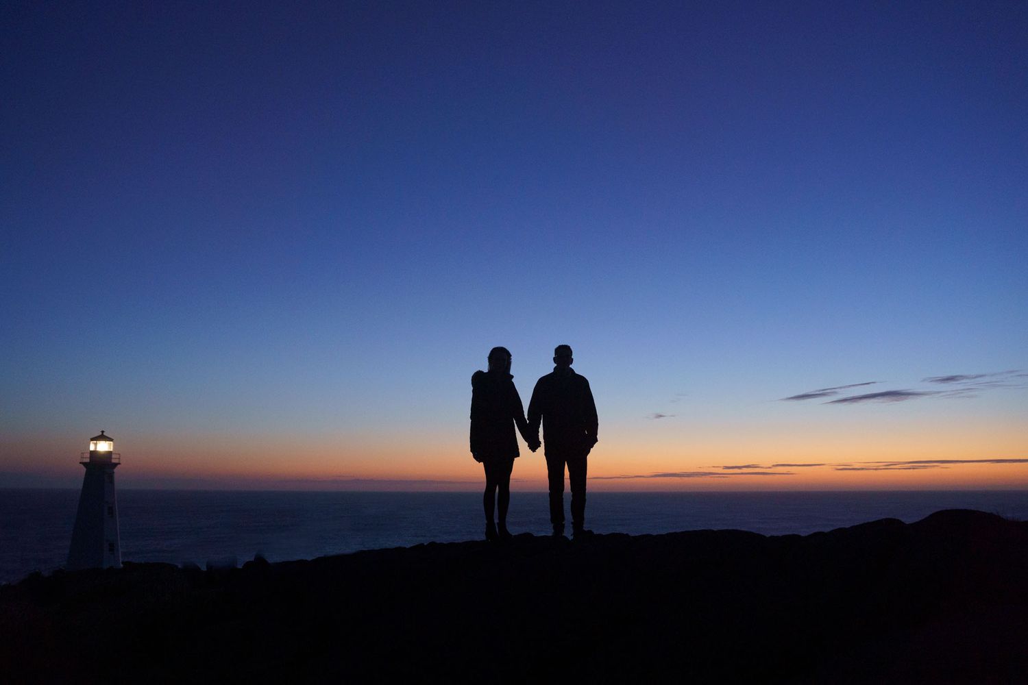 A newlywed couple standing together at Cape Spear at sunrise, with the soft glow of the morning light illuminating the rugged coastline. NL