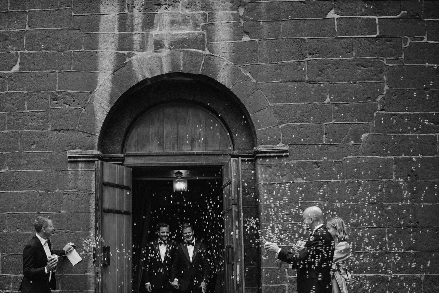 Black and white wedding photo of couple exiting church door as guests throw confetti in celebration.