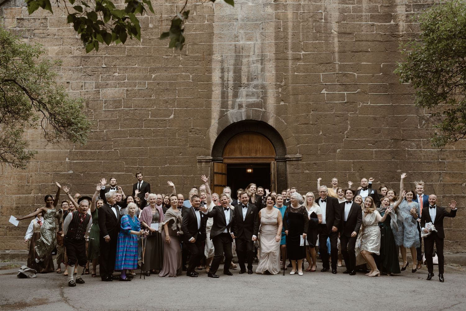 Large wedding group photo of guests celebrating outside a historic stone church with raised arms.