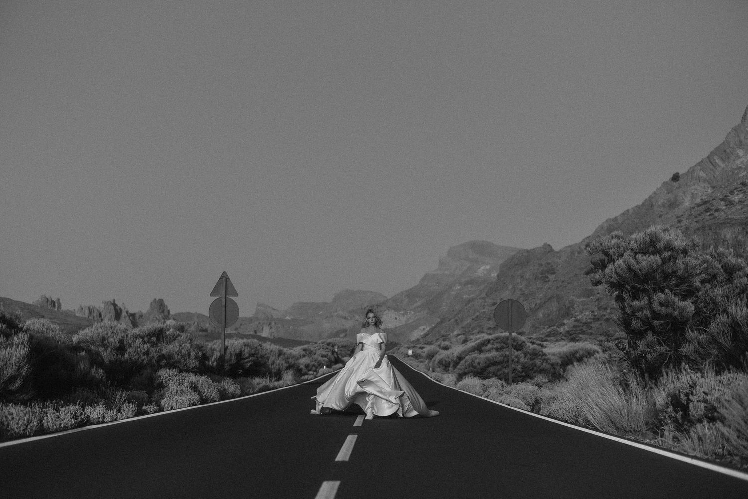 Bride in white wedding dress standing on empty road with mountains in background, black and white photo.