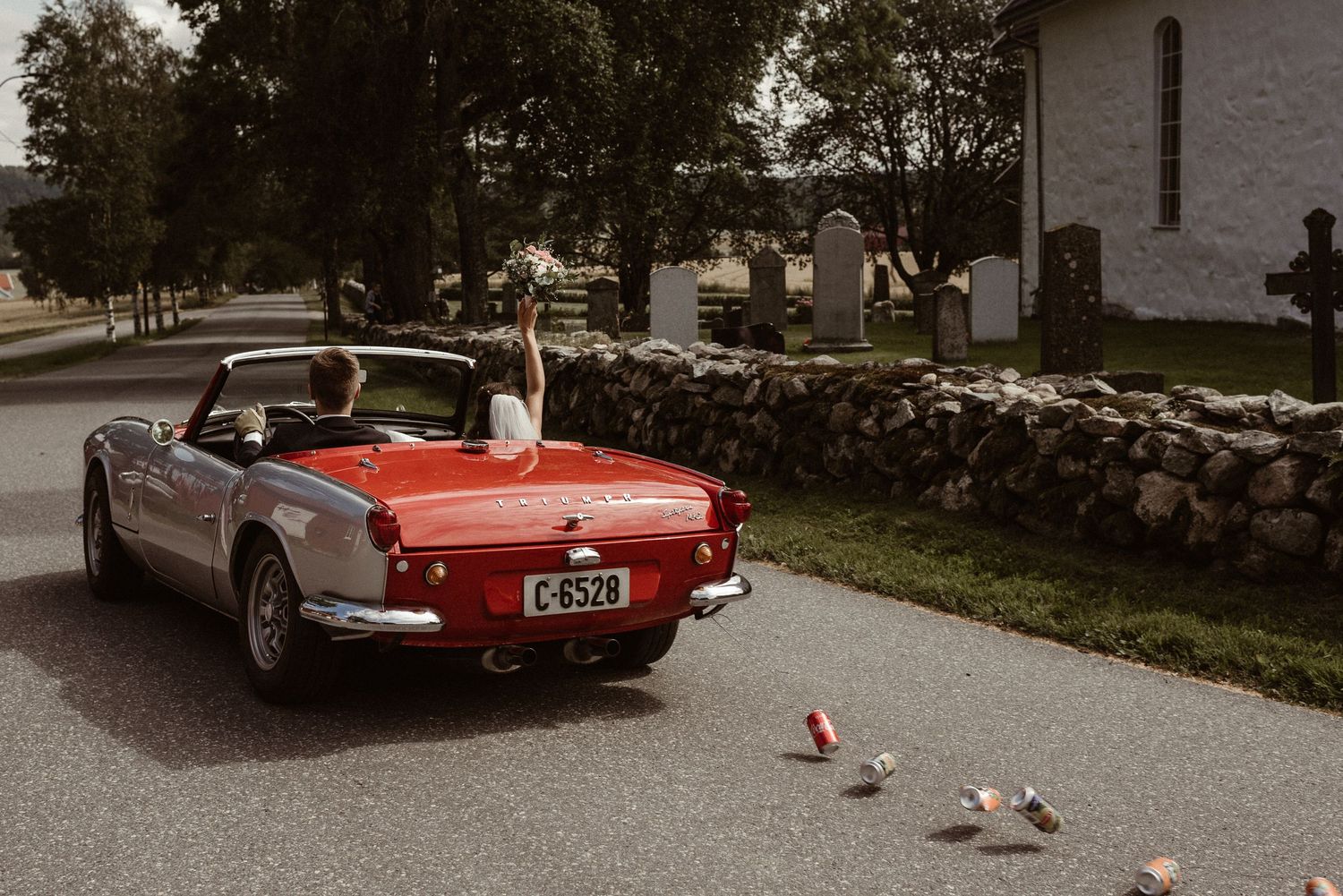 Bride and groom driving away in a classic red convertible car after their wedding ceremony.